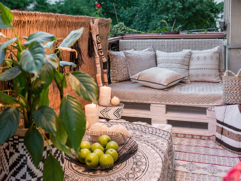 Pequeño balcón con decoración con pared de forro de bambú, un banco palet, una pequeña mesa en el centro y plantas.